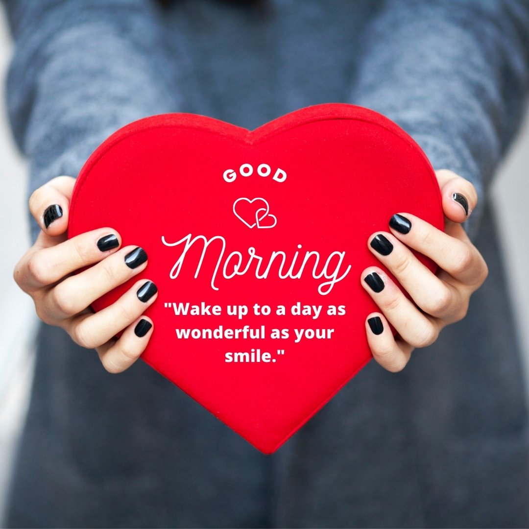 Good morning heart image featuring hands holding a large red heart with 'Good Morning' and the quote 'Wake up to a day as wonderful as your smile' on a blurred background.