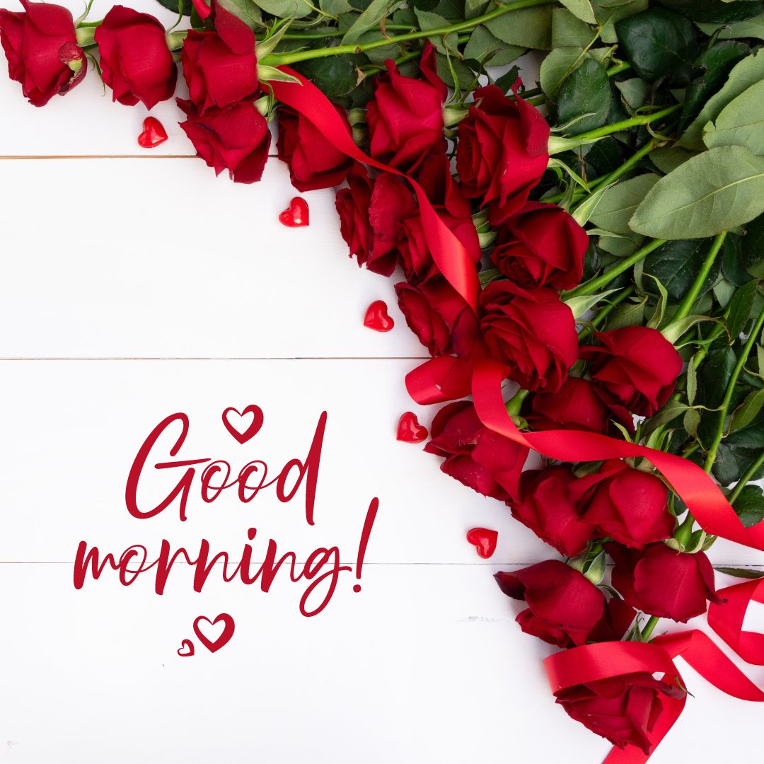 Good morning image with rose flowers featuring a cascade of lush red roses and green leaves, intertwined with red ribbons and small heart decorations, arranged on a white wooden background with the greeting 'Good morning!' in playful red script.